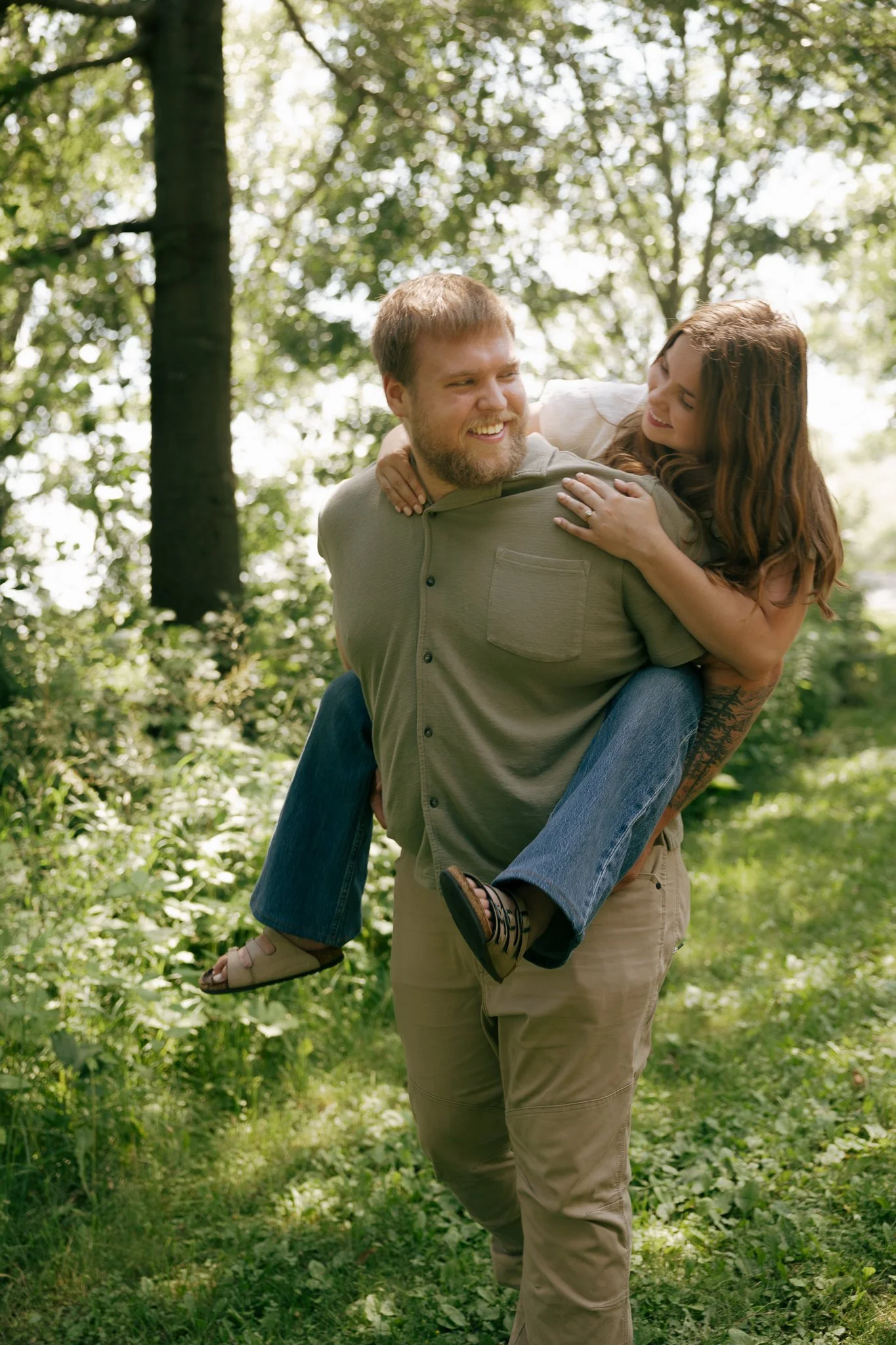 A man carrying a woman on his back in a forested area, both smiling and enjoying the moment.
