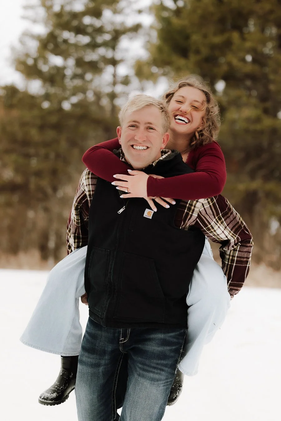 A couple enjoying winter outdoors, with the woman riding piggyback on the man's back, both smiling.