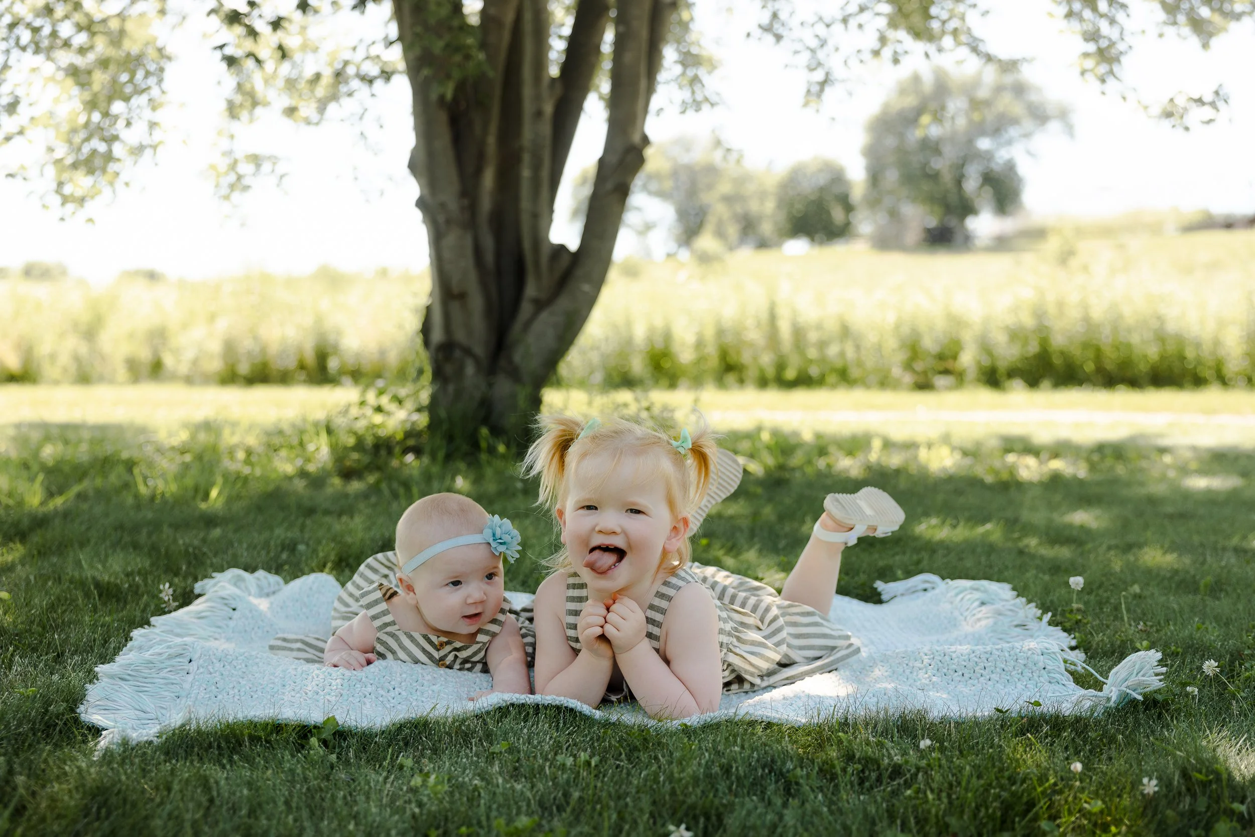 Two young children, a baby girl and a toddler girl, lying on a white blanket under a tree on a grassy field, with fields and trees in the background on a sunny day.