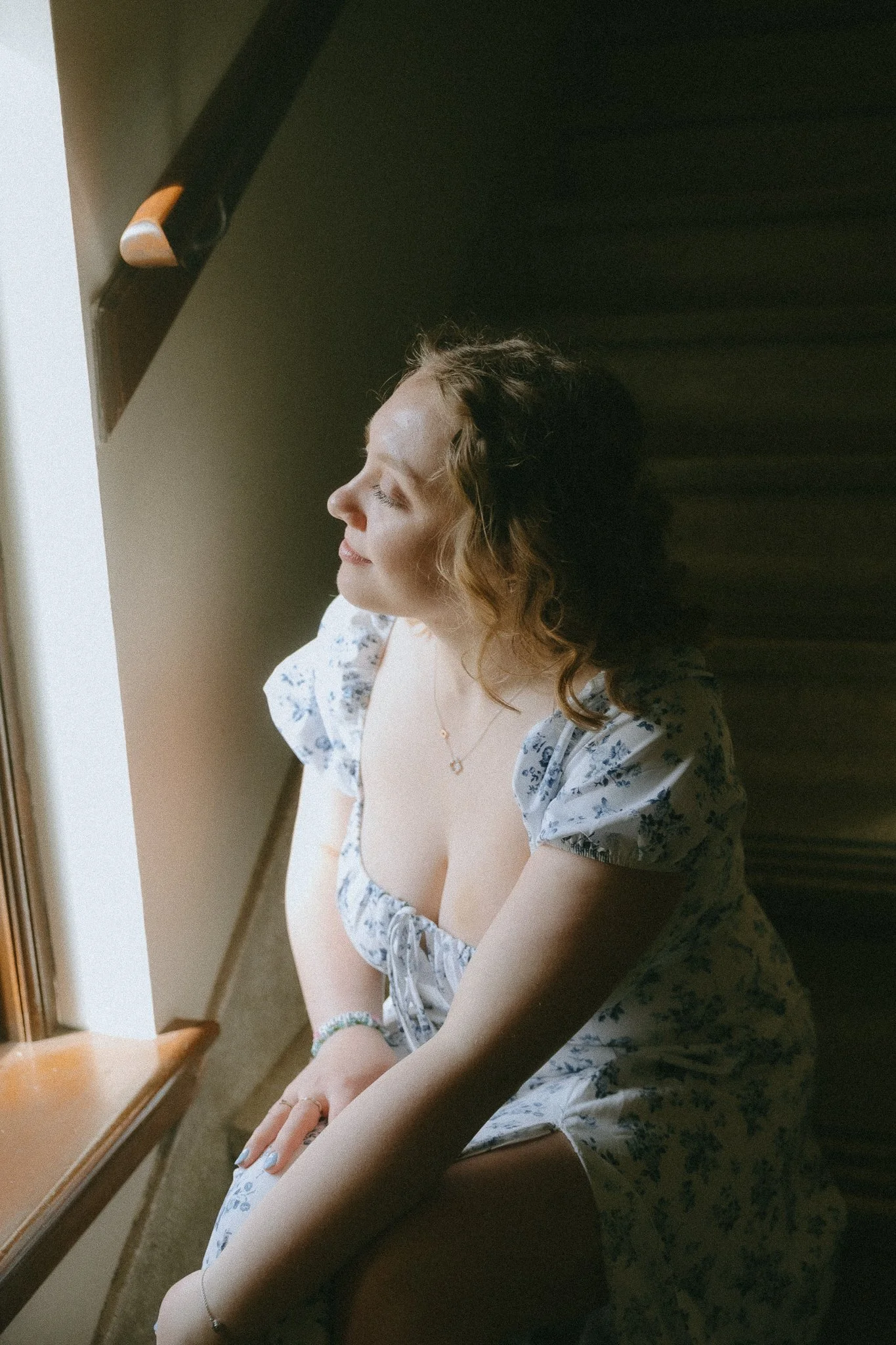 A young woman with curly hair sitting by a window, facing inward with her eyes closed, wearing a floral dress and jewelry, enjoying sunlight.