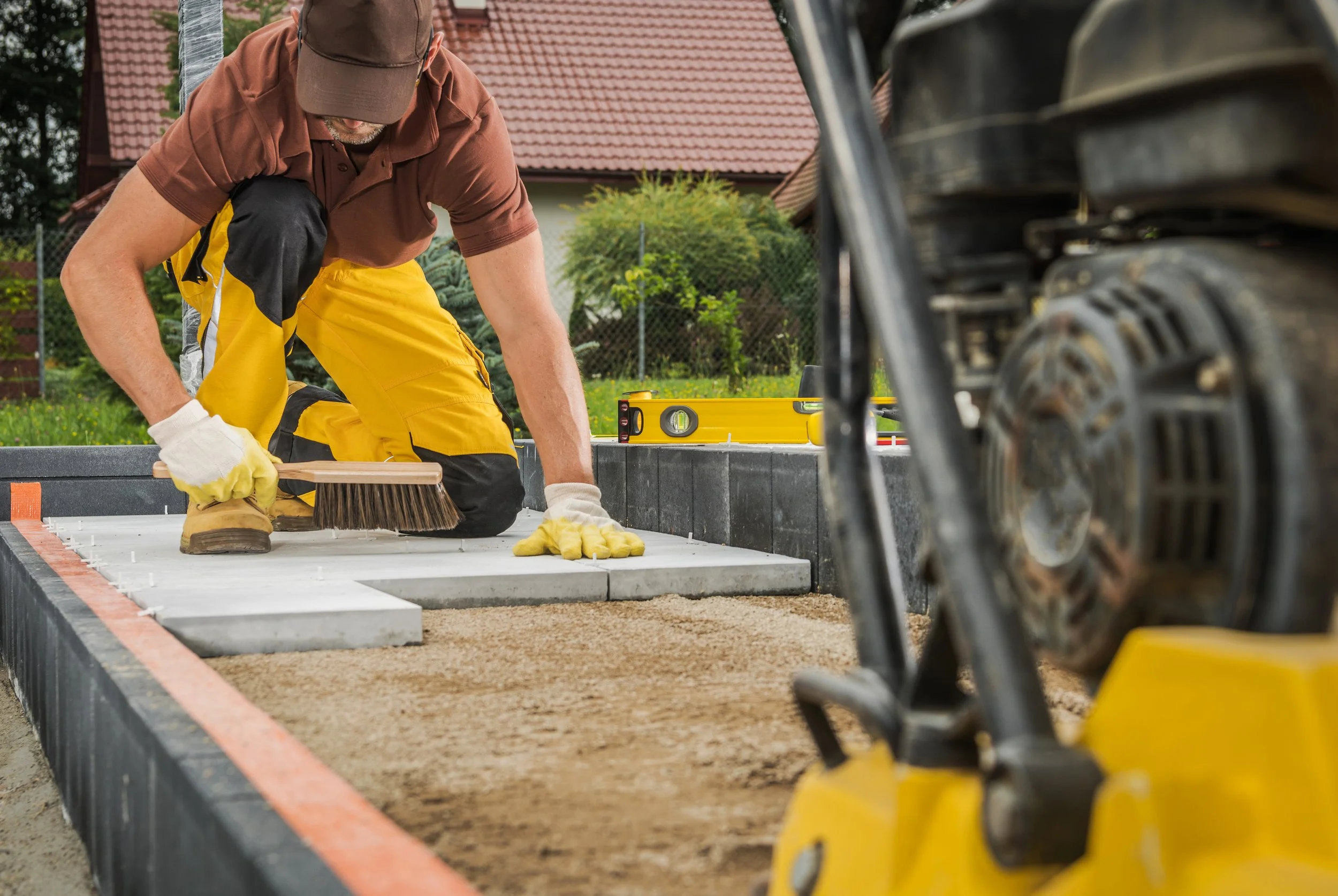 Construction worker laying paving stones and using a brush, with a compactor machine in the foreground, wearing safety gloves and a cap.