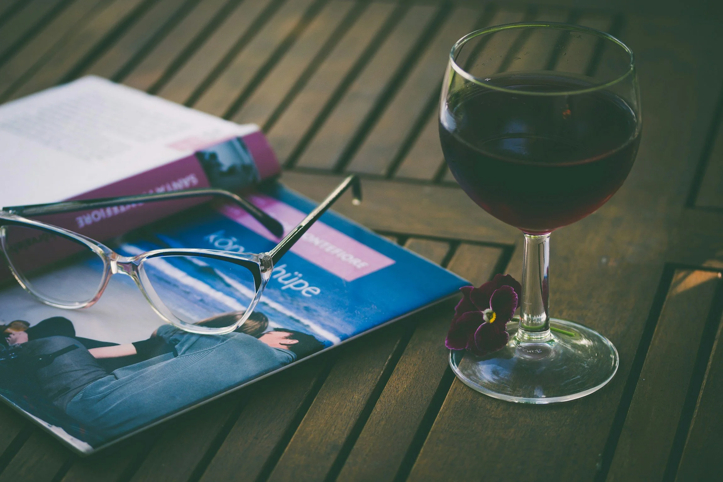A glass of red wine, a pair of clear-framed eyeglasses, and a travel magazine on a wooden table with a flower
