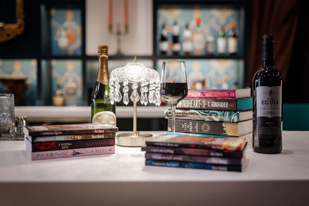 A glass of red wine, a pair of clear-framed eyeglasses, and a travel magazine on a wooden table with a flower