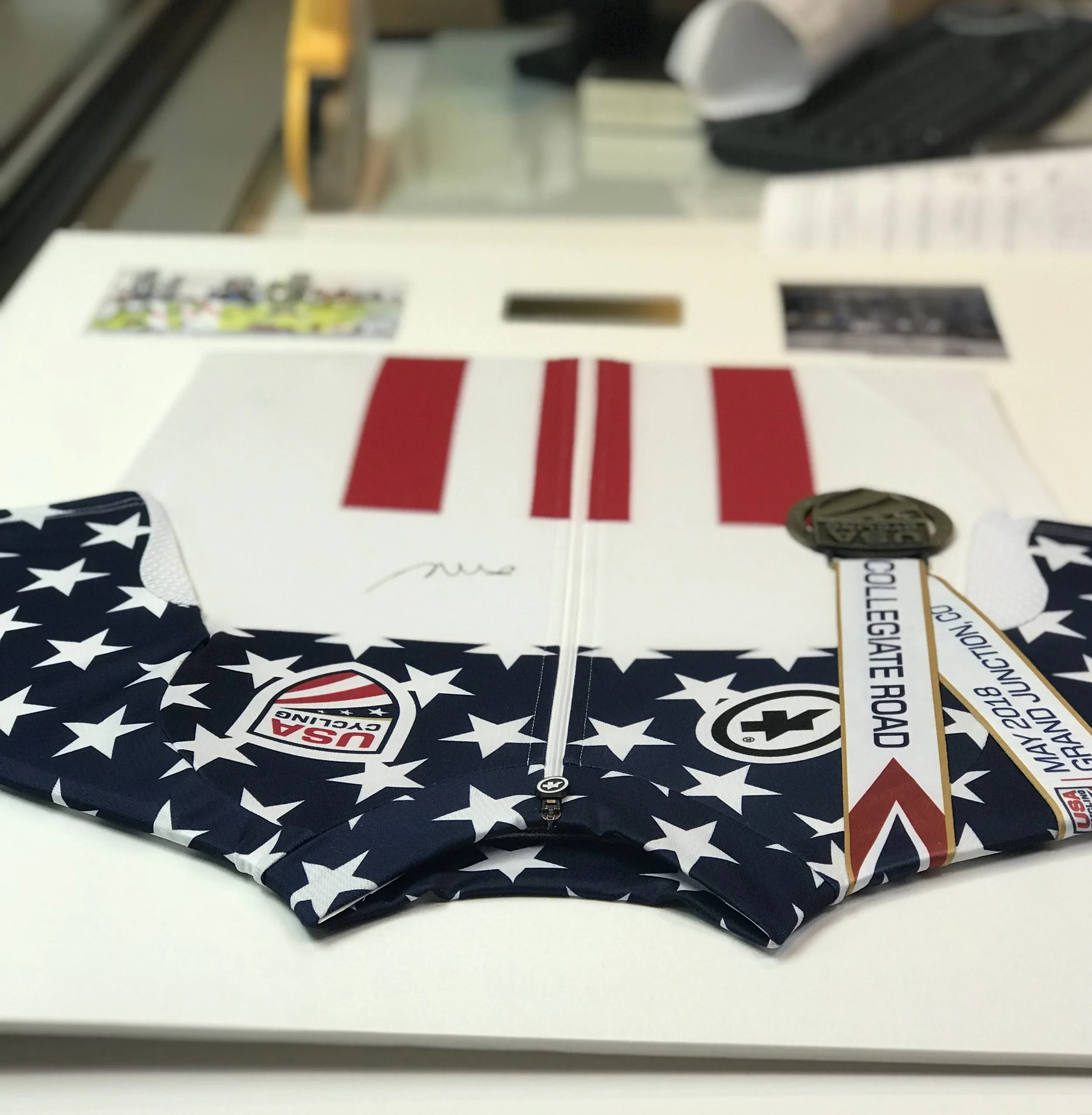 USA Cycling jersey with stars and stripes design, featuring a medal labeled 'Collegiate Road,' on a table with other papers and objects.
