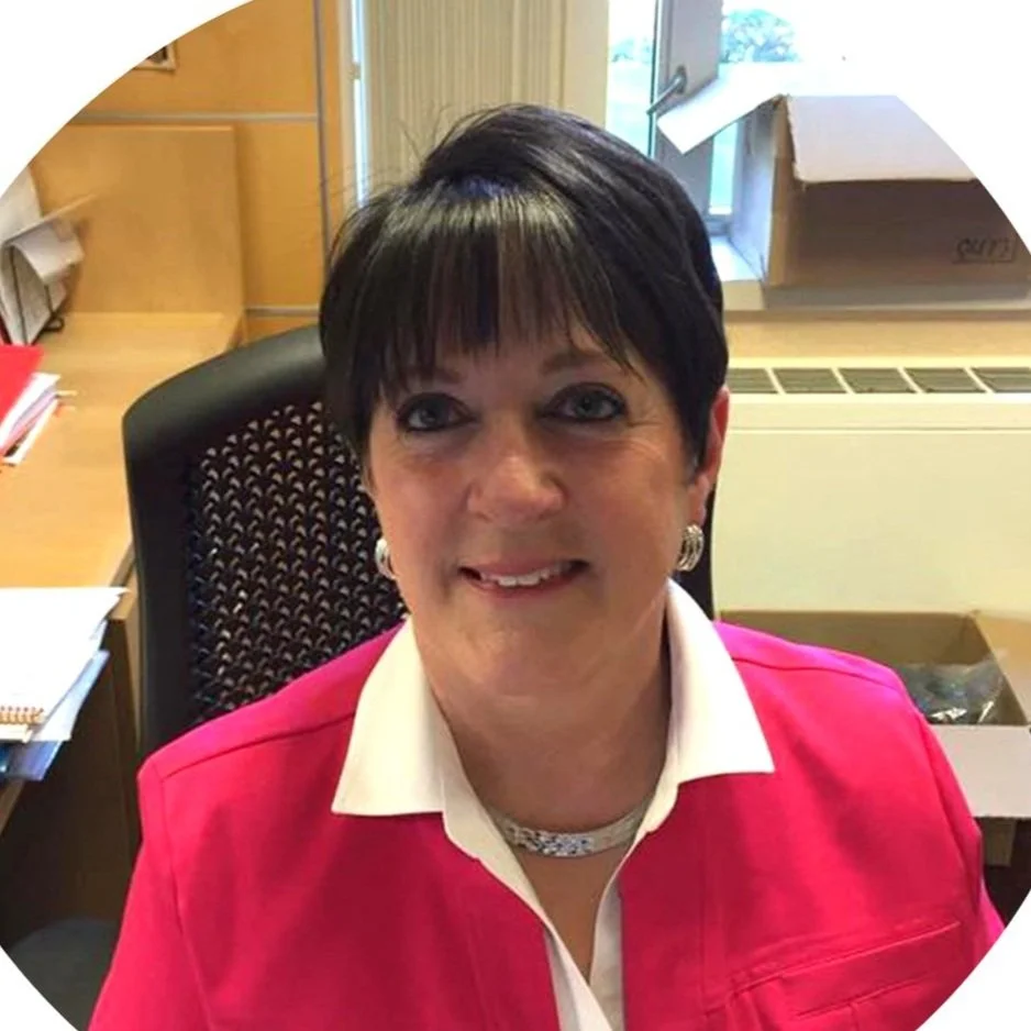Smiling woman with short dark hair wearing a pink jacket in an office setting