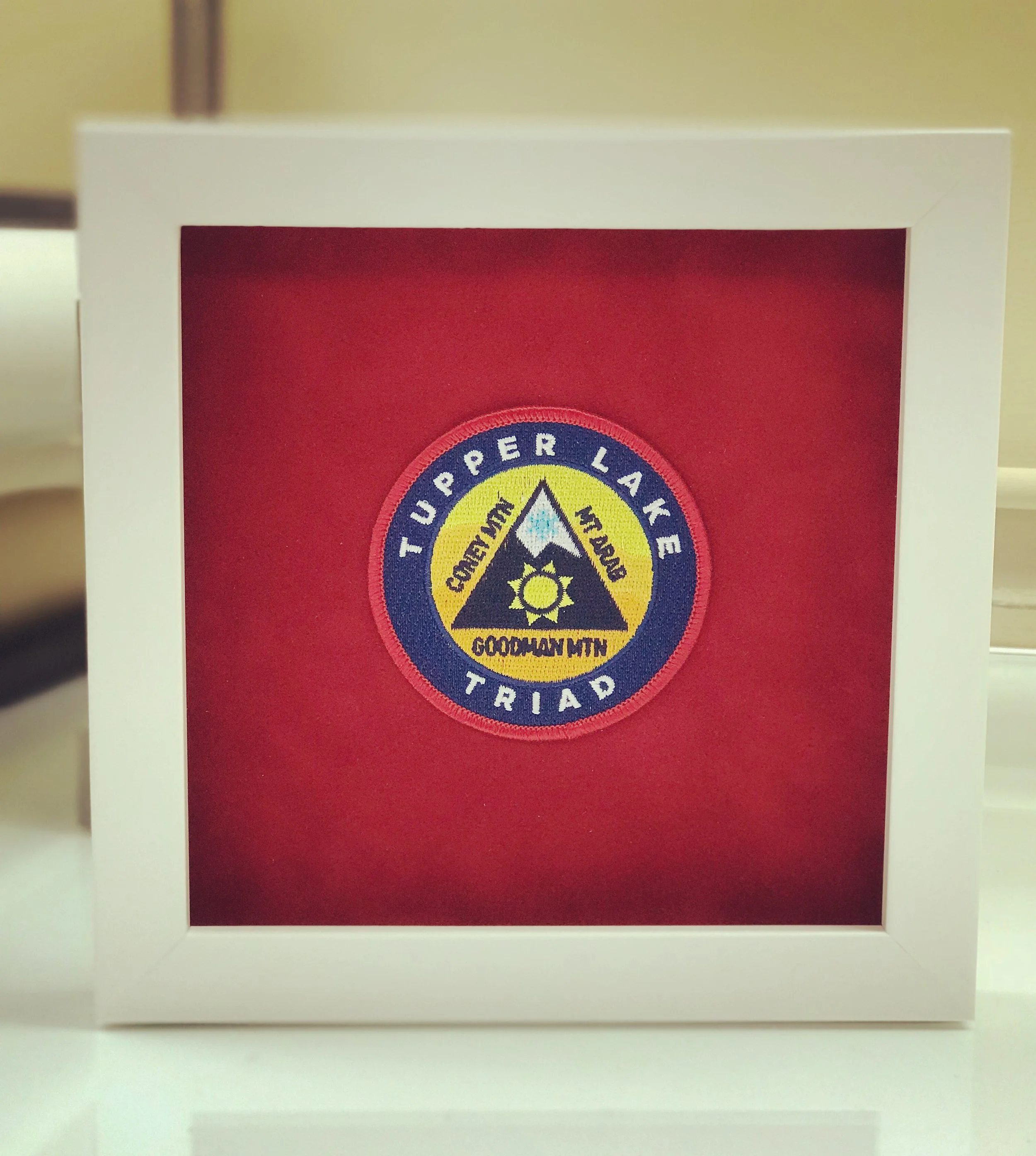 Tupper Lake Triad patch framed in a white border with red background, featuring names Coney Mountain, Goodman Mountain, Mt. Arab, and a sun design.