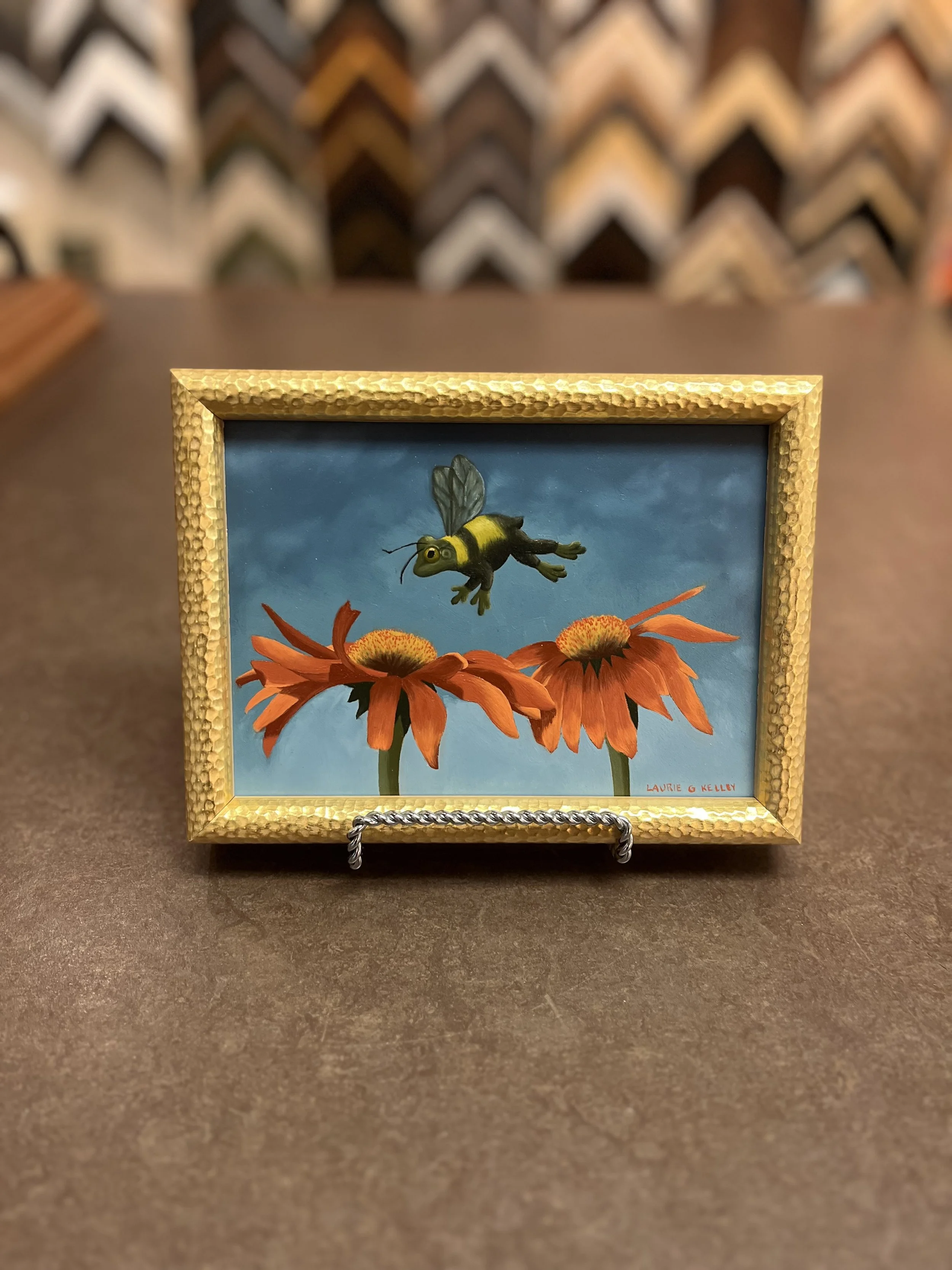 Framed painting of a cartoon bee hovering over two orange flowers, displayed on a stand.