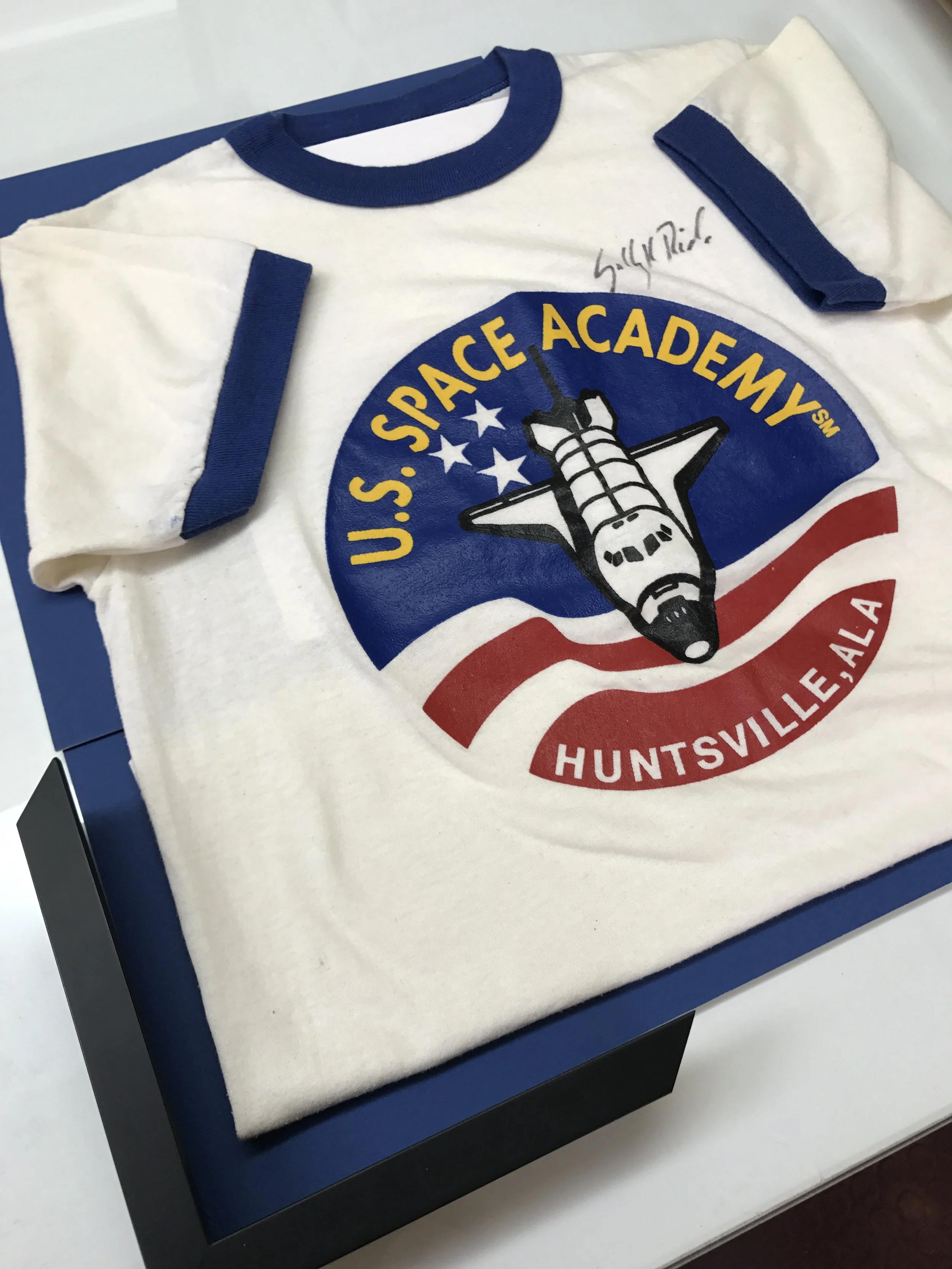 A white T-shirt with a blue collar and sleeve ends featuring the logo of U.S. Space Academy in Huntsville, Alabama, displaying a space shuttle and American flag elements. The shirt is autographed.