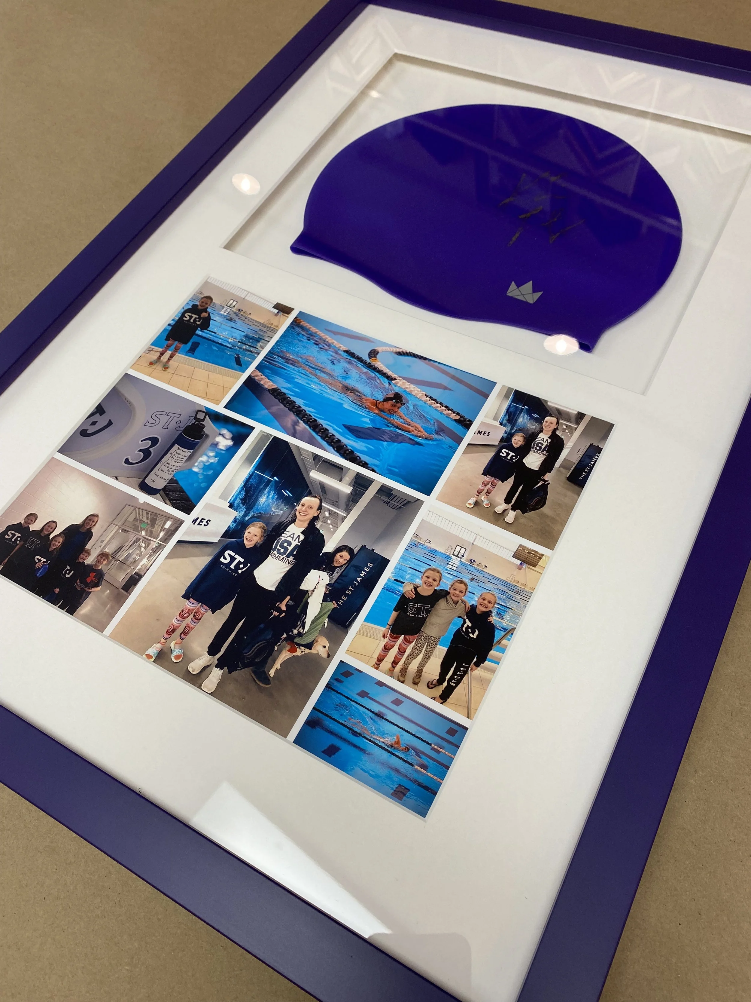 A framed collage featuring a purple swim cap and multiple photos of children and swimmers participating in swimming activities. The frame has a purple border, and the photos show people in a swimming pool and on the pool deck. Some children wear matching outfits and there appears to be a swimming event or team setting.
