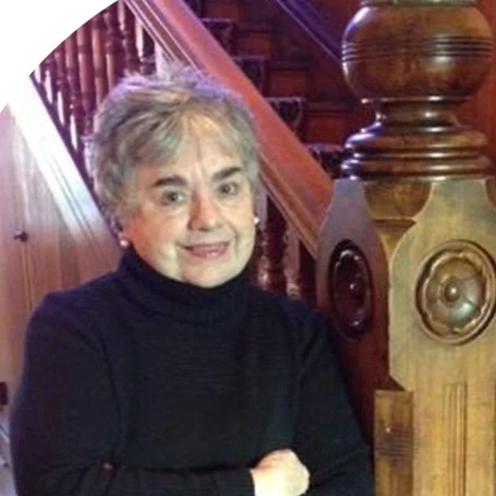 Elderly woman standing by a wooden staircase with ornate design, wearing a black turtleneck sweater.