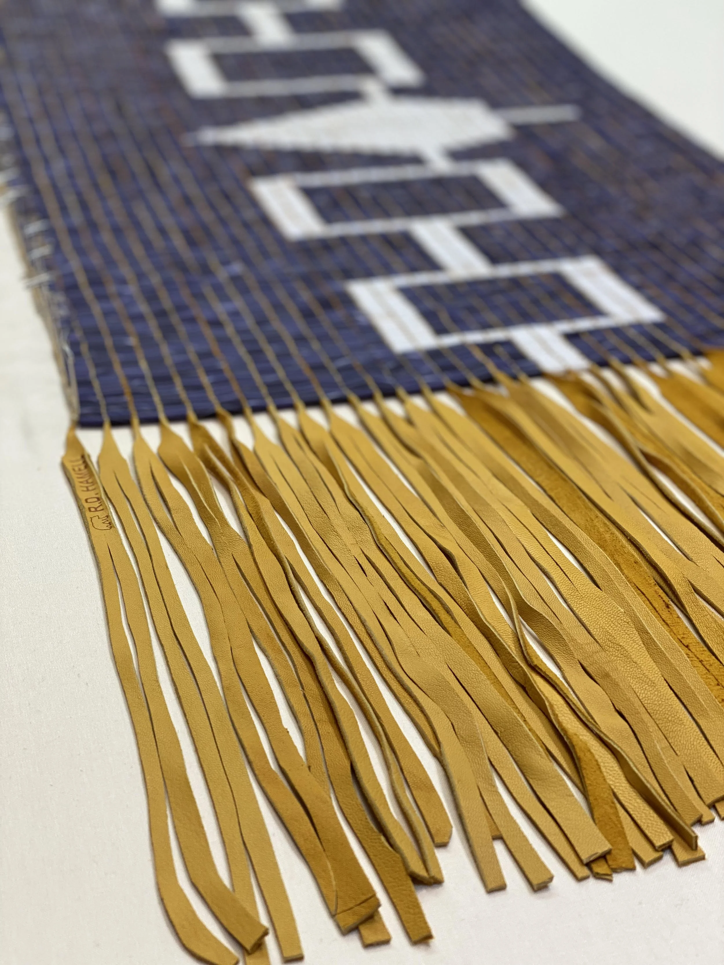 Close-up of a woven textile with yellow fringes on one end. The fabric has blue threads with white geometric patterns.