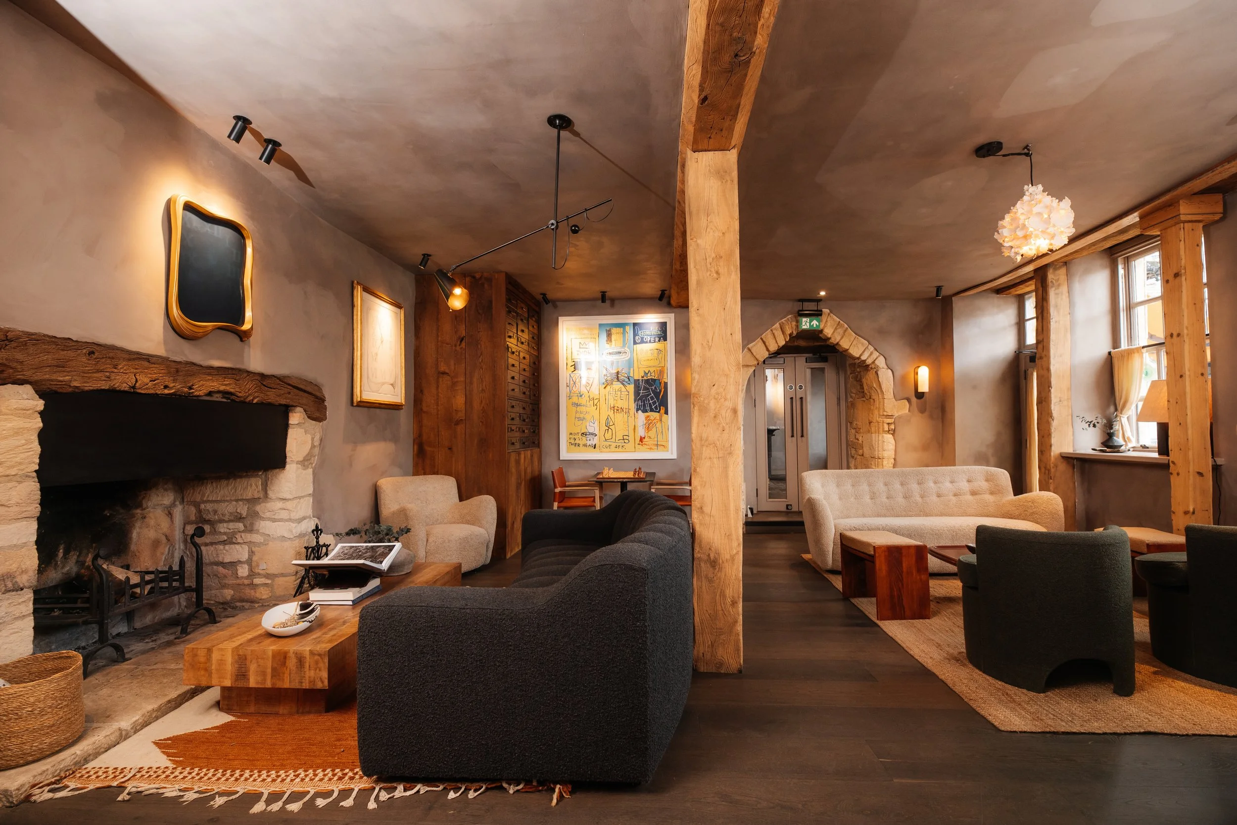 A cozy living room with a rustic design, featuring a wood-beamed ceiling, a stone fireplace, various framed artwork on the walls, and several comfortable sofas and chairs. There are large windows with curtains natural light, and decorative lighting fixtures hang from the ceiling.