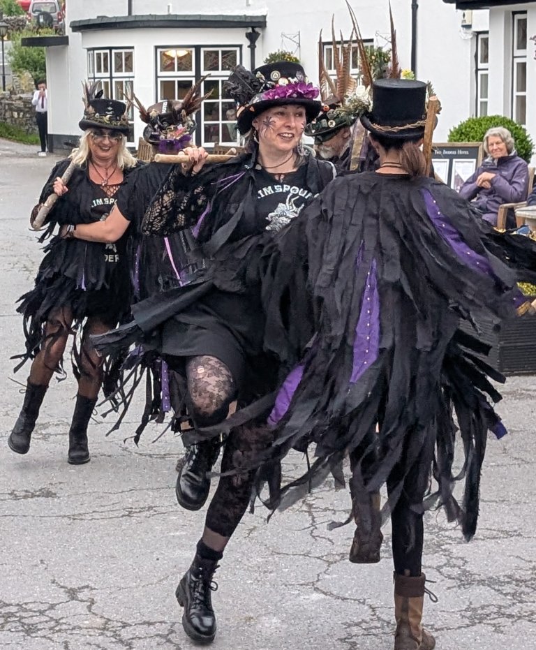 Group of women dressed in black, purple, and feathered costumes, wearing hats with feathers and decorations, participating in a parade or festival on a street.
