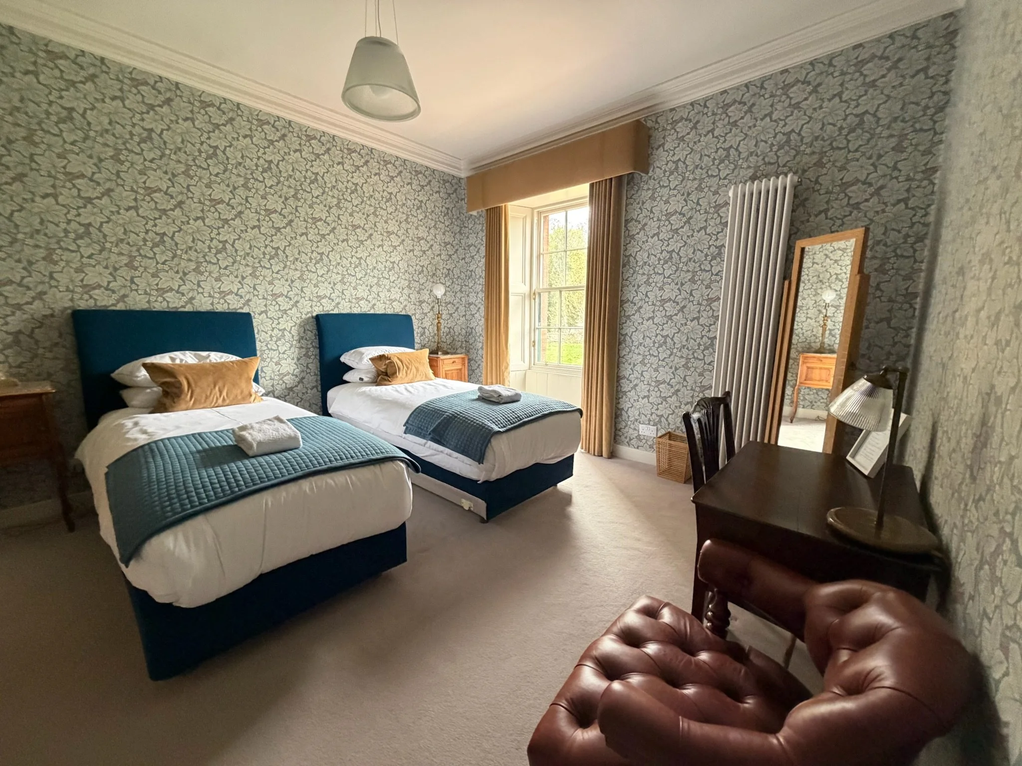 A bedroom with two beds that have blue headboards and white bedding, gold and beige pillows, a wooden nightstand with a lamp, a large window with beige curtains, a mirror leaning against the wall, and a radiator next to the window.