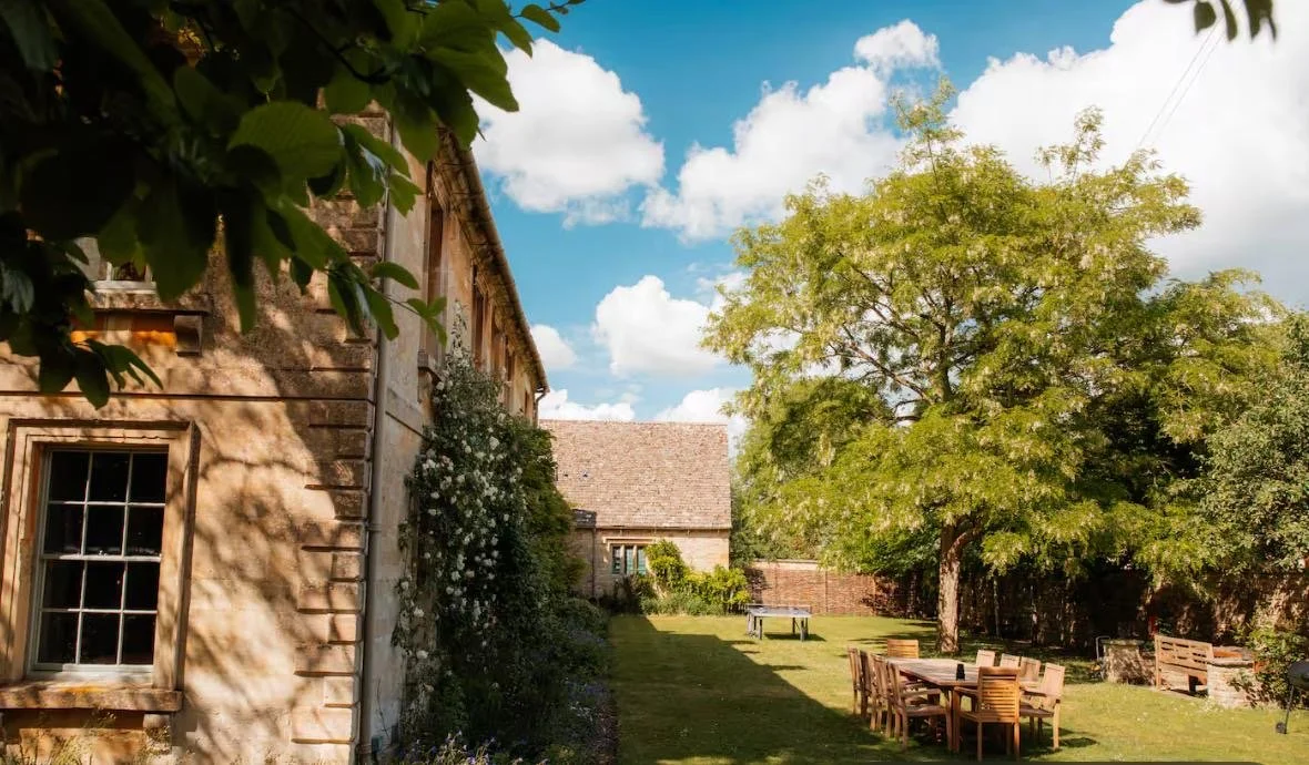 A backyard garden with a stone house on the left, surrounded by greenery, a large tree, a wooden dining table with chairs, and a sky with fluffy clouds.