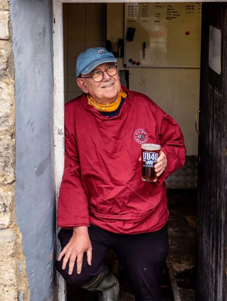Older man with glasses and a blue cap, wearing a red jacket and yellow bandana, holding a pint of beer, smiling while standing in a doorway.