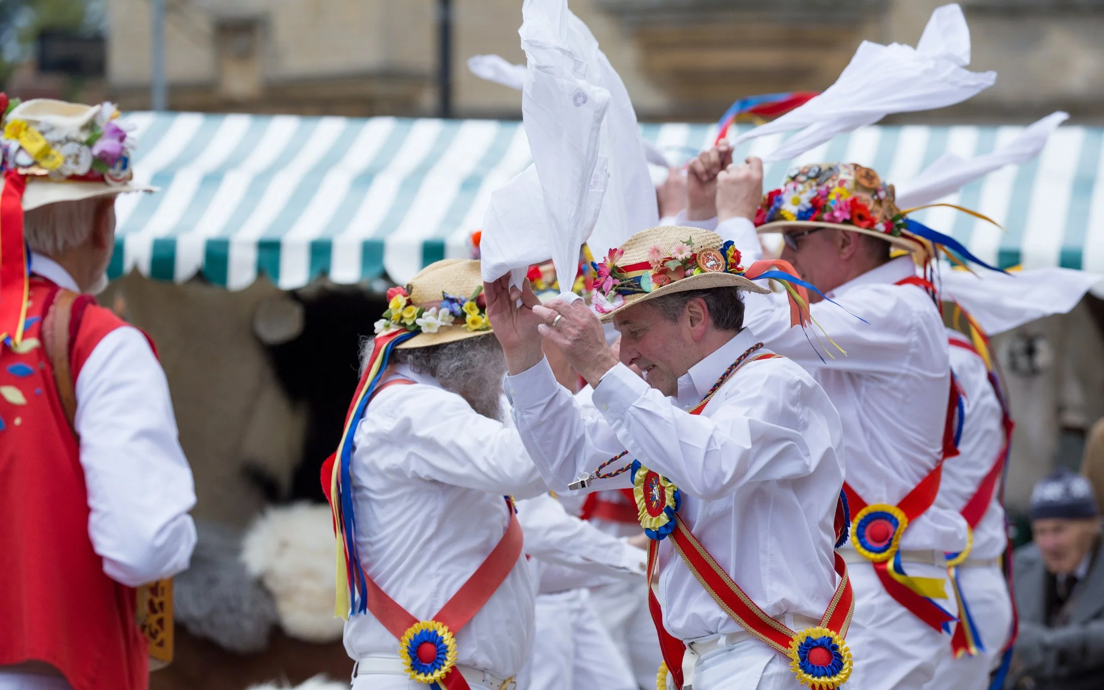 Group of people dressed in traditional folk costumes, dancing or performing at an outdoor event, with colorful ribbons and floral hats.