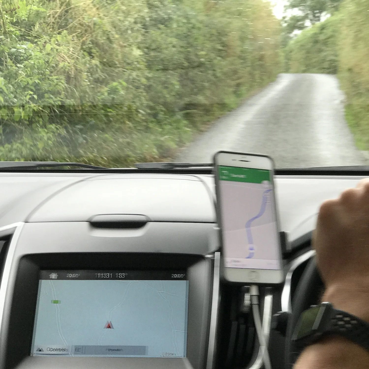 Interior car view of a narrow backcountry road in Great Britain, with hedges lining the road and GPS navigation visible on a mounted phone and dashboard screen.