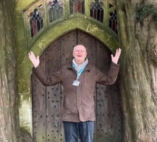 A man smiling and waving standing in front of a wooden door with stained glass windows, surrounded by a tree.