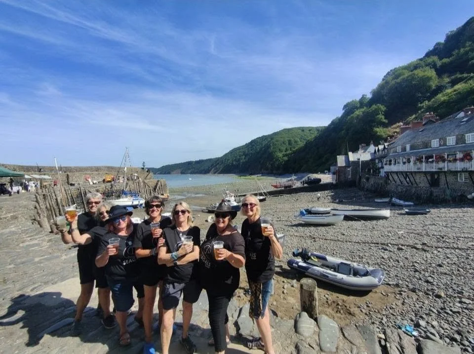 A group of seven people, mostly women, standing together on a rocky beach with boats and a hillside in the background, enjoying drinks on a sunny day.