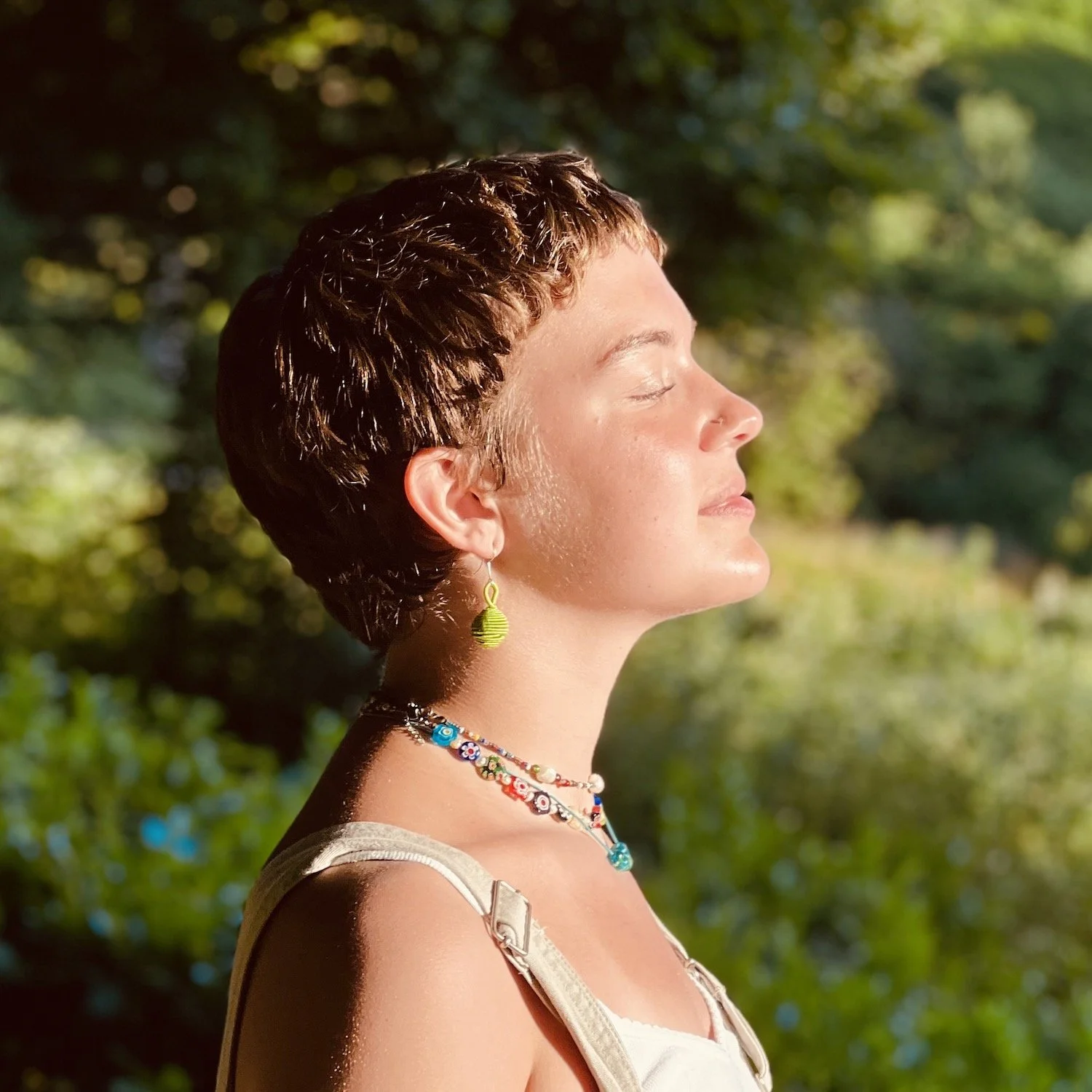 A person with short hair and colorful jewelry basks in golden sunlight in a wooded area, eyes closed in peaceful reflection. This serene moment captures the wellness and nature-focused experiences shaping current UK travel trends.