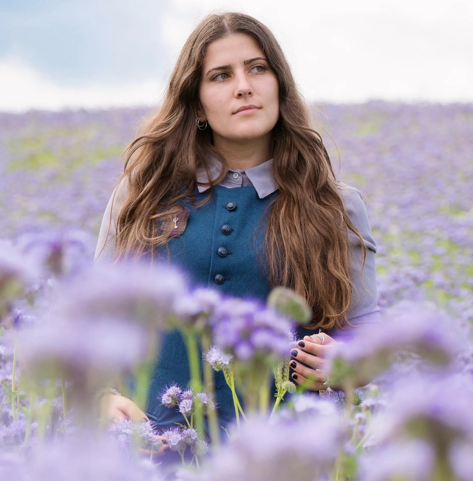 A woman with long brown hair and fair skin standing in a field of purple flowers, wearing a blue vest with black buttons and small hoop earrings, looking to the side.
