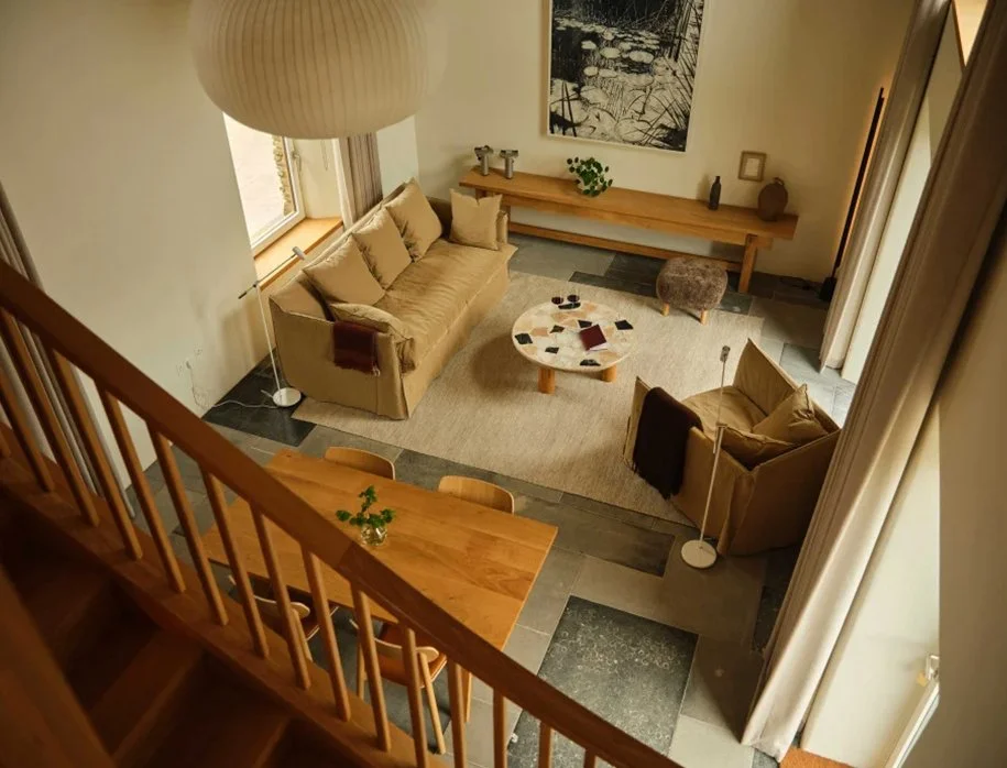 A cozy living room viewed from the staircase, with a beige sofa and armchair, a round coffee table with items on it, a wooden sideboard with decorative objects, a large black-and-white artwork, a tall floor lamp, and curtains on the windows.