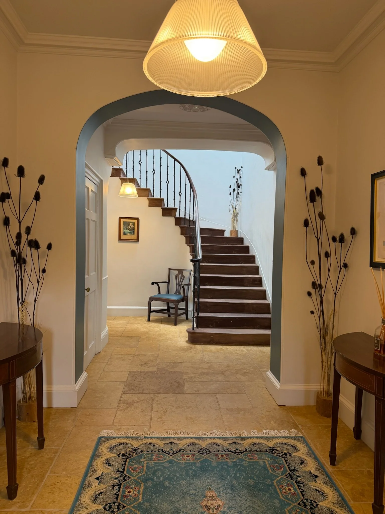 The image shows a well-lit hallway with a large ceiling lamp, a staircase with wooden steps and a metal railing, a small chair, and decorative plants. The floor features a patterned rug, and the walls are adorned with framed pictures and dried floral arrangements.