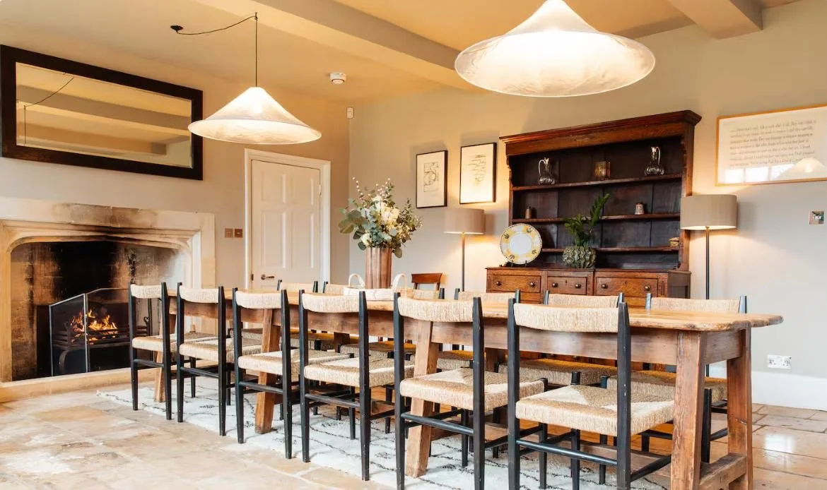A dining room with a large wooden table surrounded by ten matching chairs with woven seats. There are two large pendant lights hanging from the ceiling, a fireplace with a metal guard on one side, a dark wooden cabinet with decorative items, framed artwork on the wall, and a large vase with flowers on the table.