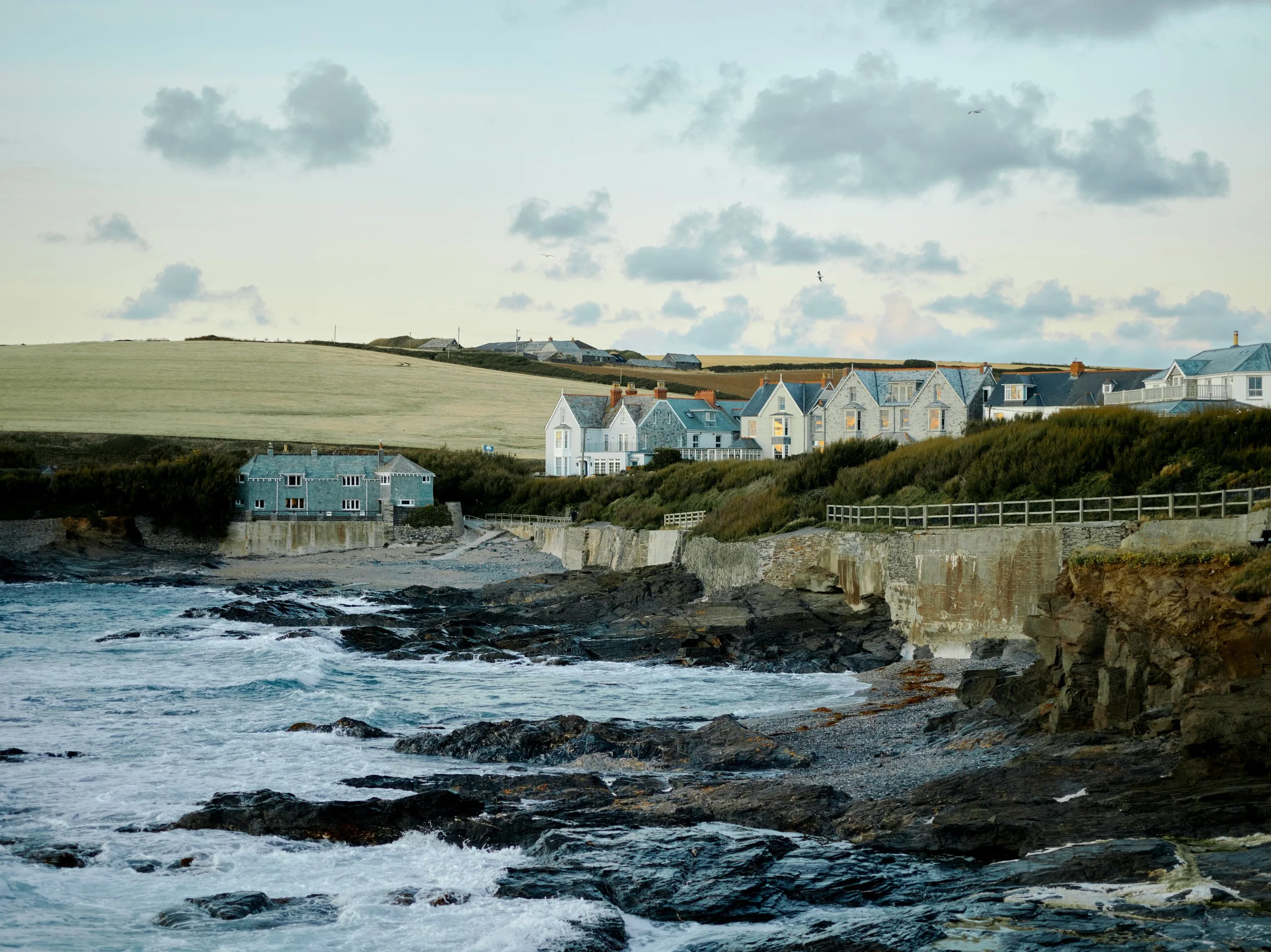 View of a coastal neighborhood with rocky shoreline, houses, grassy fields, and cloudy sky.