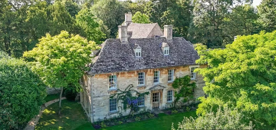 A historic stone house surrounded by lush green trees and a well-maintained lawn, with a sunny sky.