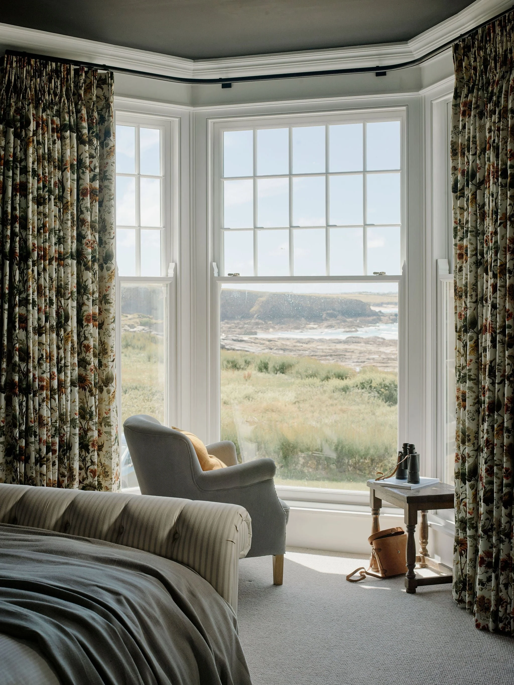 A cozy bedroom with a large bay window with floral curtains, a white armchair with a yellow pillow, a small table with binoculars and a brown leather bag, and a bed with a striped blanket. Outside the window, there is a scenic view of a coastline with rocks and water.
