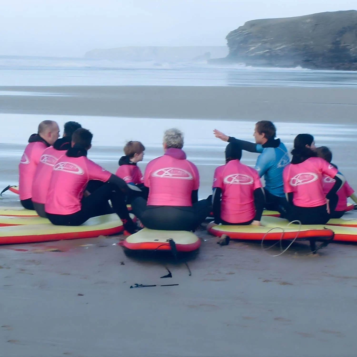 A surf instructor in a blue wetsuit leads a group lesson on a misty beach, with students in pink wetsuits seated on surfboards. Surfing lessons like this are increasingly popular, especially along coastal destinations shaping UK travel trends.