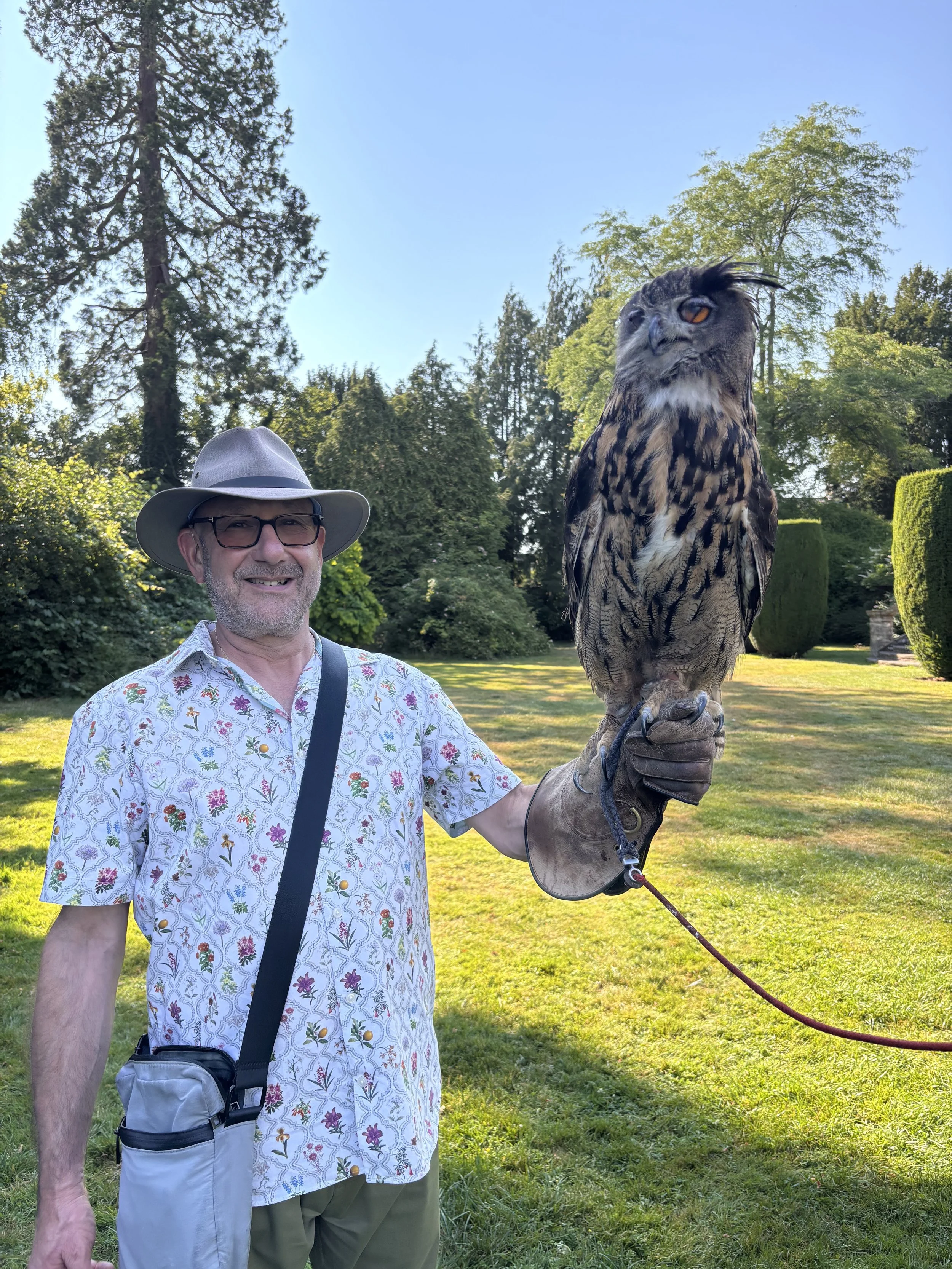 A man wearing glasses, a floral shirt, and a wide-brimmed hat holding a large owl with a glove on its talons in a park with trees and a clear blue sky.