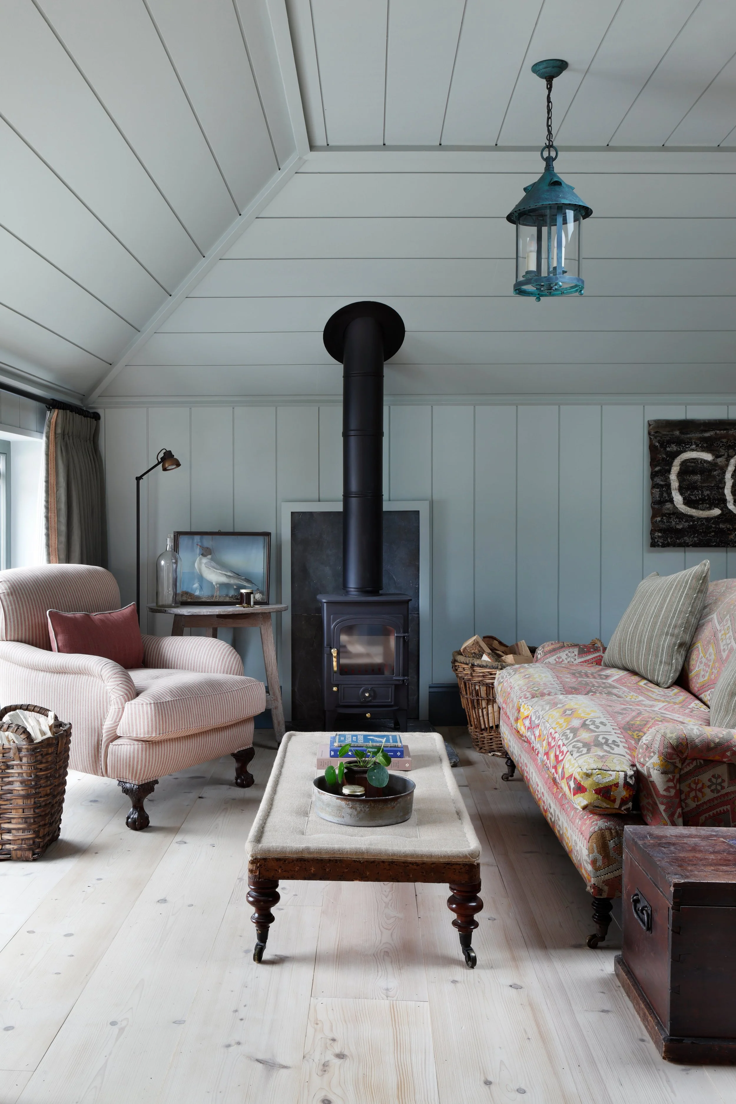 Living room with light wooden floor, pink-striped armchair, floral couch, wood stove with black chimney, coffee table with books, potted plant, decorative items, framed bird picture, and hanging blue lantern light
