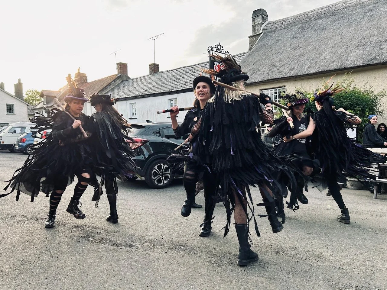 Group of women dressed in black costumes with feathers and hat accessories, performing a dance outdoors on a street.