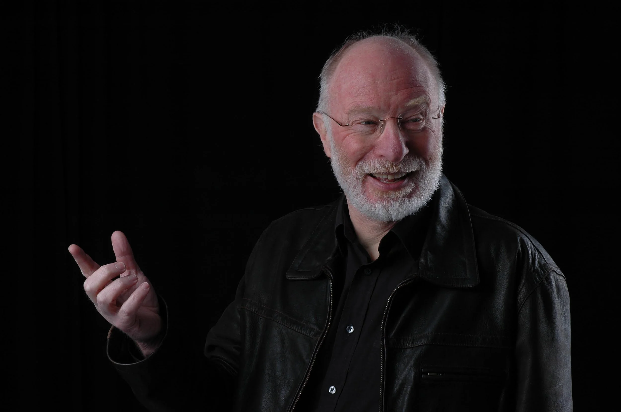 An elderly man with a white beard and glasses is smiling and looking to his left. He is making a peace sign with his right hand and is dressed in a black leather jacket and black shirt against a black background.