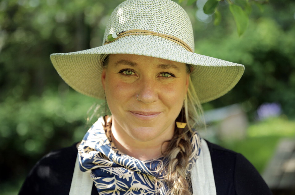 Woman wearing a wide-brimmed straw hat, a blue and white patterned scarf, and a black shirt outdoors with green foliage in the background.