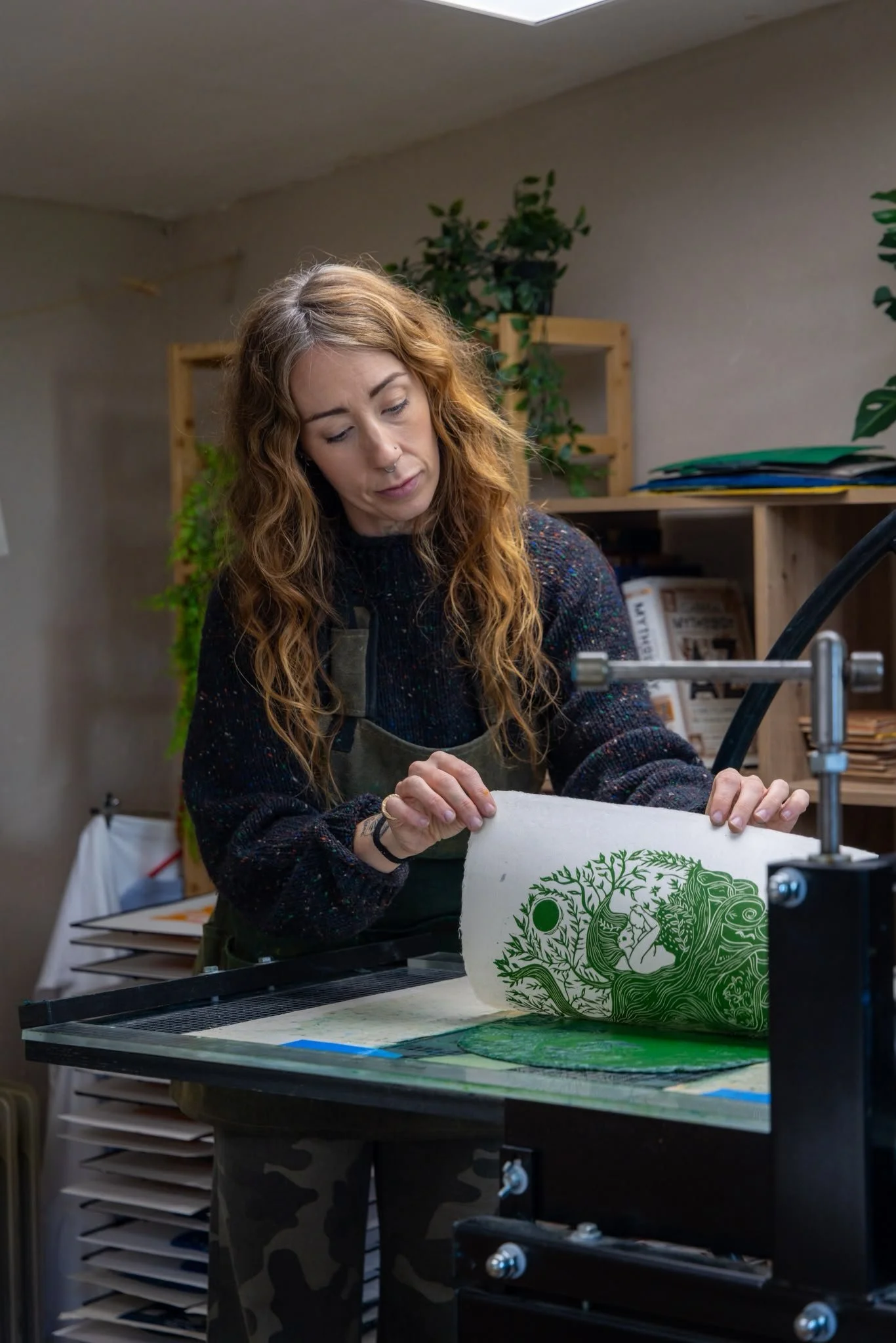 A woman with curly reddish hair and wearing a green apron, working on a print project using a printing press in a workspace with plants and shelves in the background.