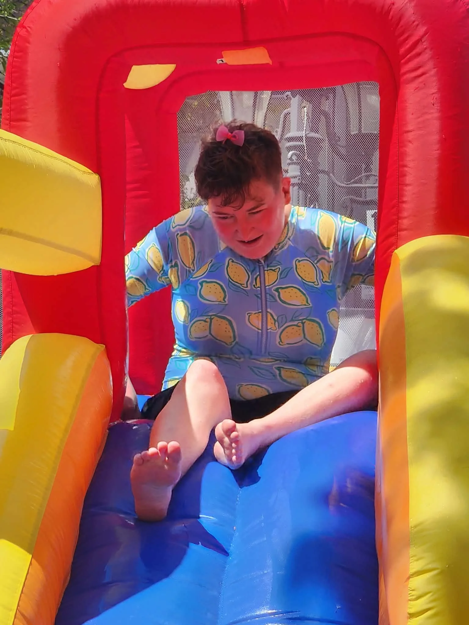 Child with a pink bow in their hair sitting inside a colorful inflatable bounce house, with one arm holding their knee and the other hand resting on their foot.