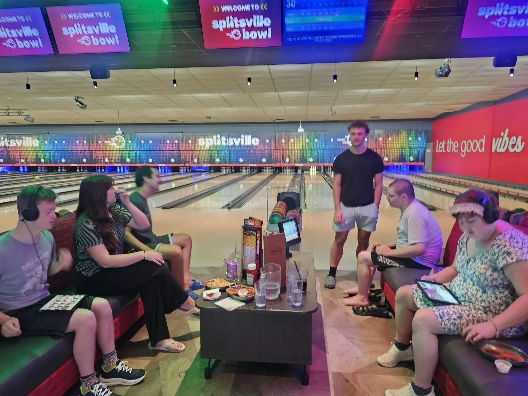 People sitting and standing in a lively indoor bowling alley, with multiple colorful electronic screens displaying scores and advertisements, and a group of friends enjoying food and drinks.
