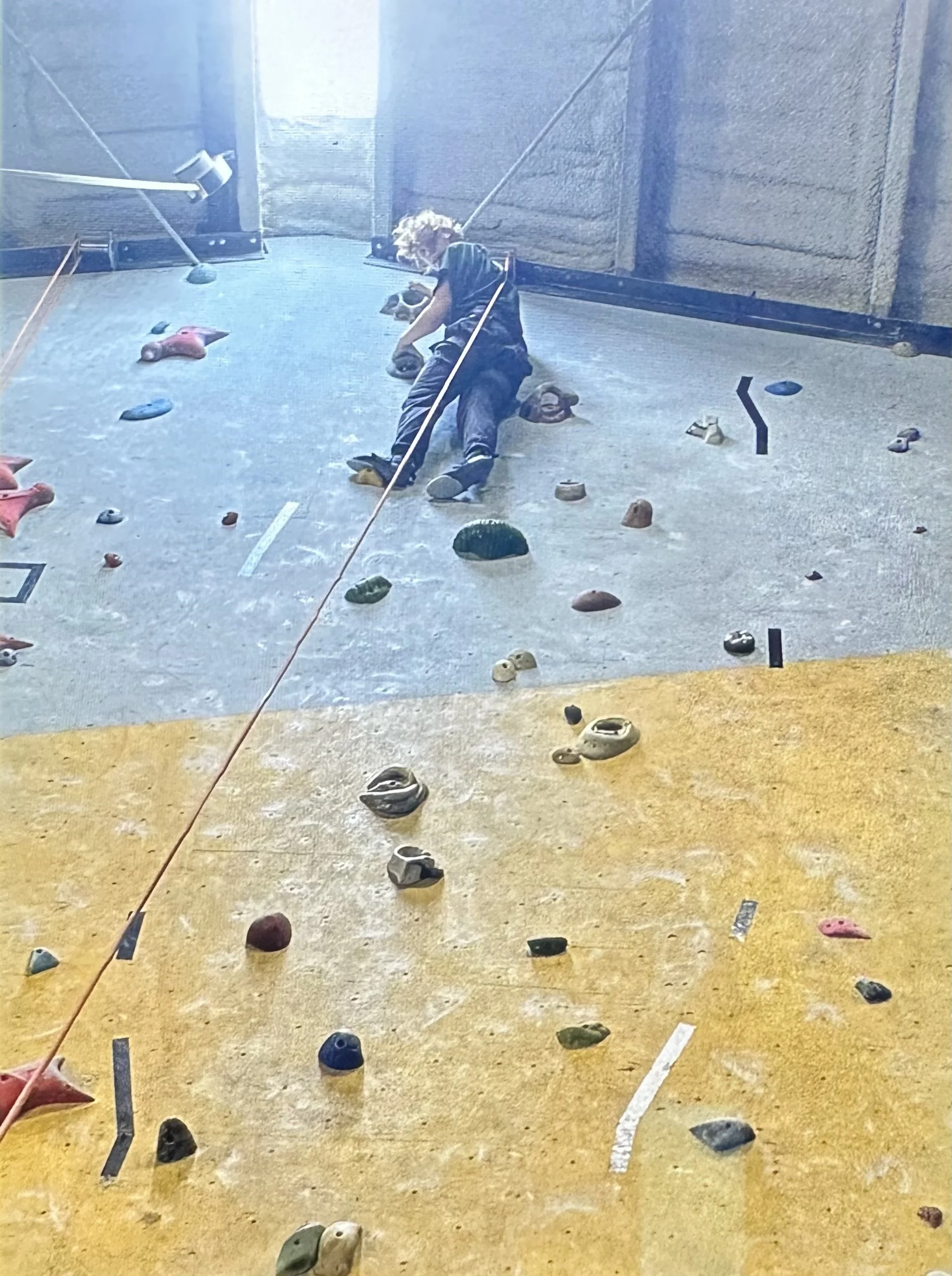 A person with curly hair climbing an indoor rock wall with various holds and a safety rope.