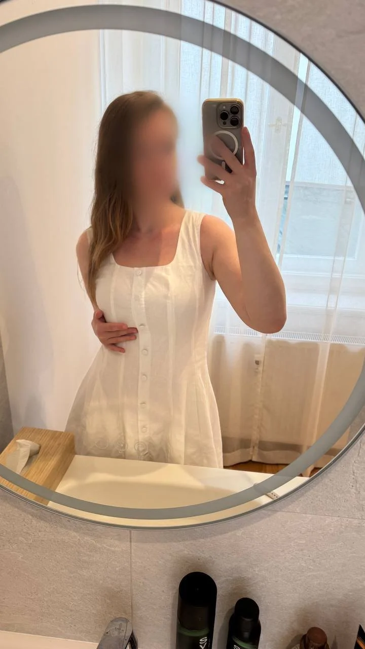 Veronica taking a mirror selfie in a room with curtains, wearing a cream sleeveless dress, with her face blurred for privacy.
