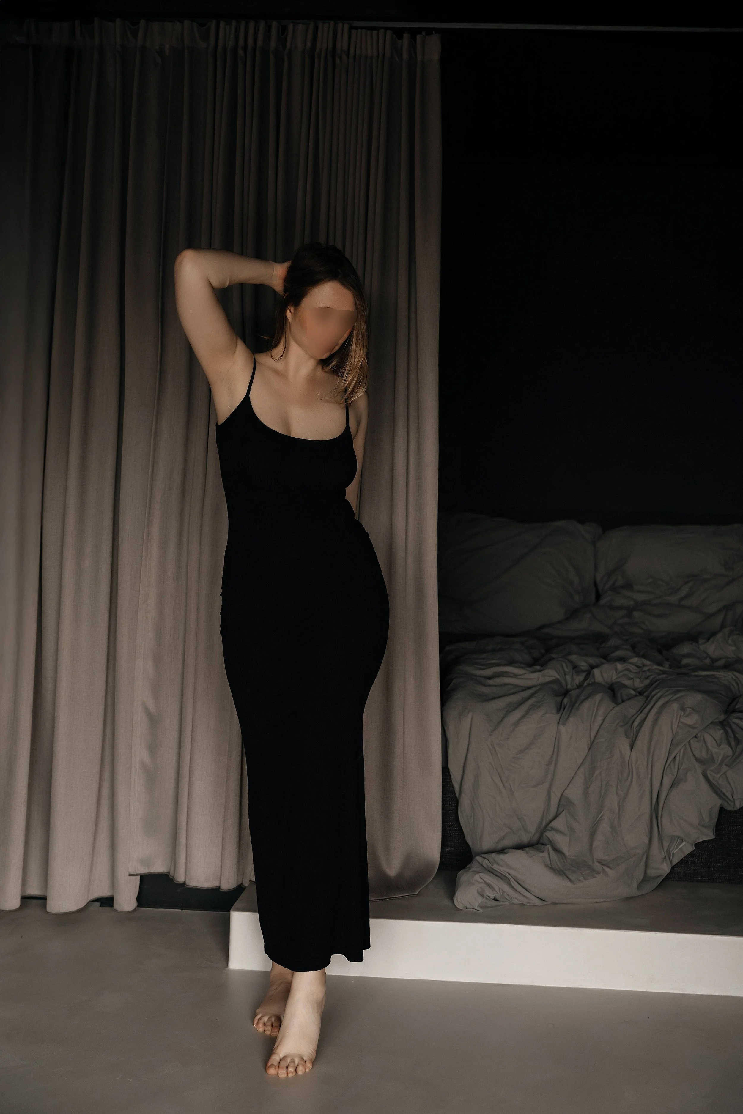 Veronica Ellis is standing by the bed, wearing an elegant black dress, in a luxurious loft