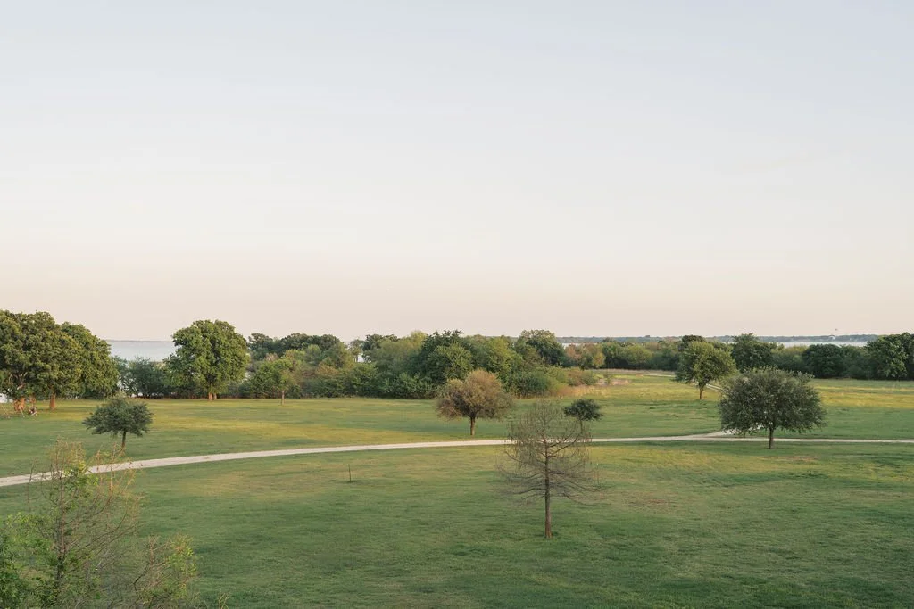 A scenic landscape with a grassy field, several trees, and a winding pathway under a clear sky at Peninsula Ranch in DFW 