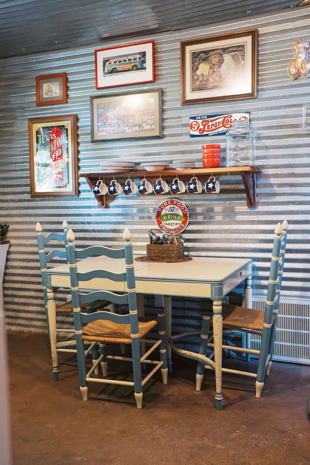 A cozy dining area with a white table and four blue chairs with woven seats, against a corrugated metal wall decorated with vintage-style framed posters, a wooden shelf holding ceramics and a glass pitcher at Peninsula Ranch in DFW 