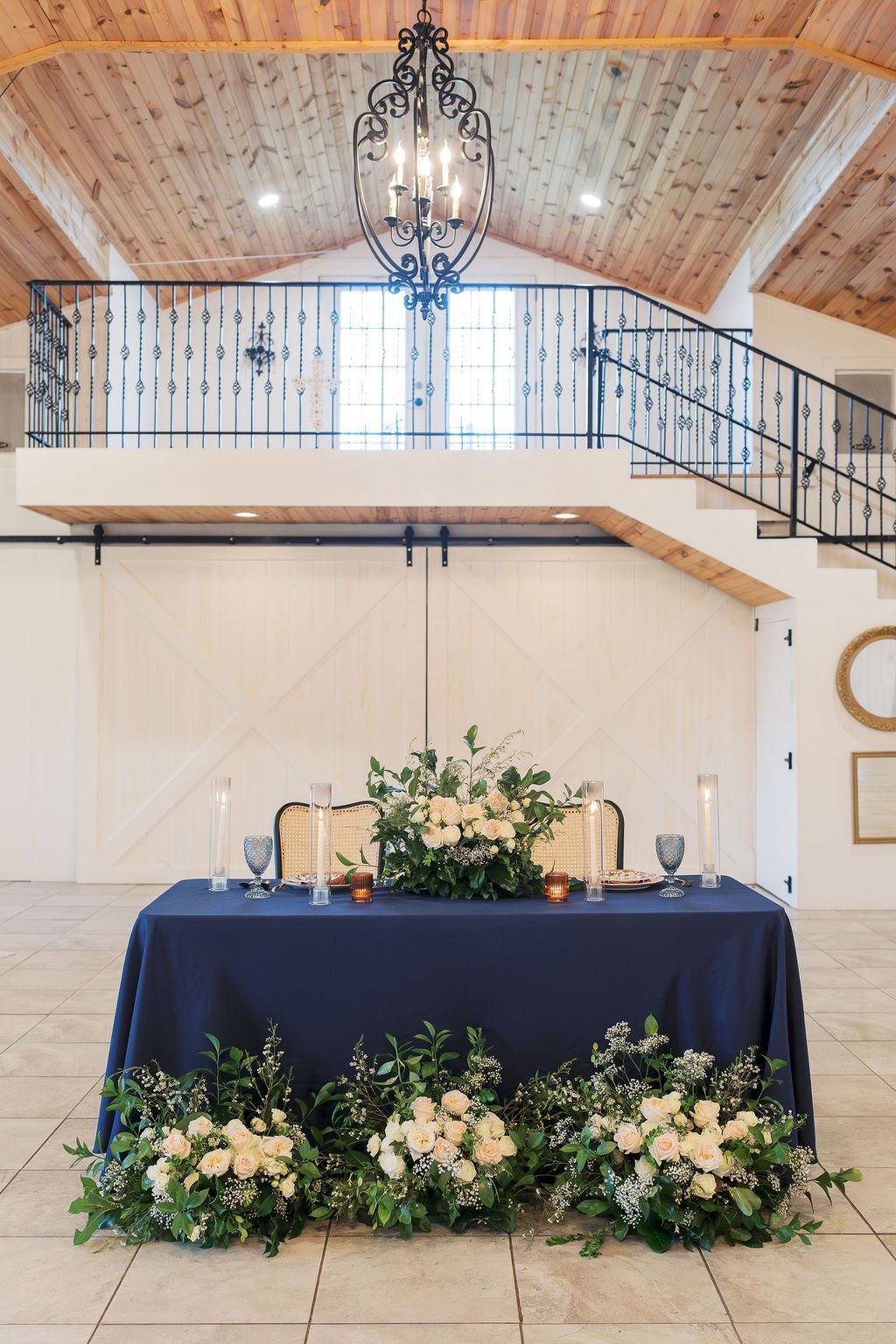 Wedding reception table with IZALENA Floral House centerpiece set in front of a white barn door, inside a venue with wood-paneled ceiling and metal staircase with decorative railing at Peninsula Ranch in DFW.