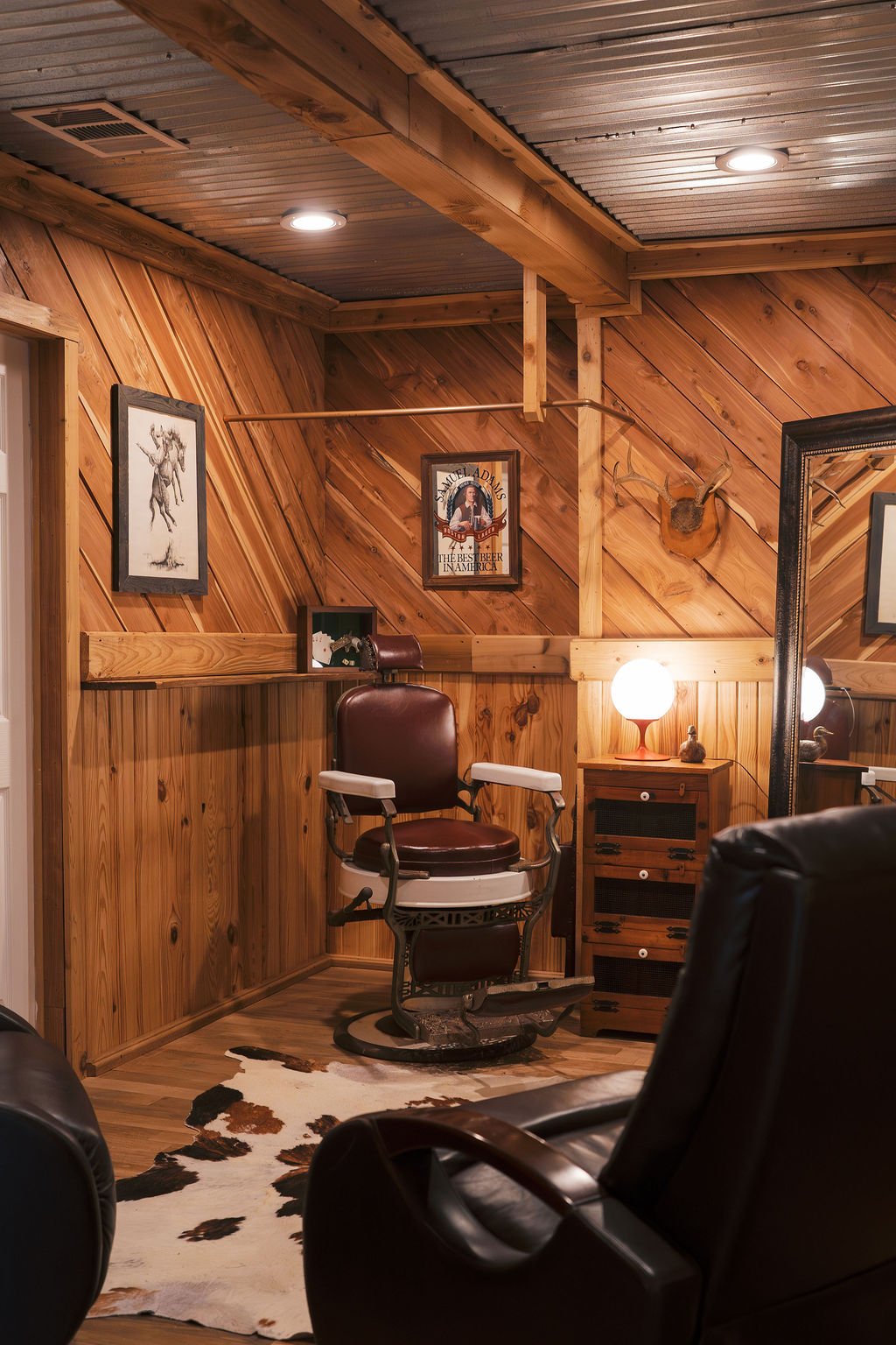 A vintage barber shop-style chair in our Groom Get Ready Room with wood-paneled walls and ceiling, decorated with framed art and a mounted deer head, with a cowhide rug on the floor at Peninsula Ranch in DFW.