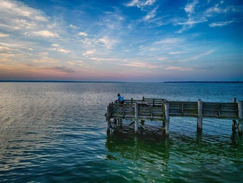 A person sitting on a wooden dock on Lake Tawakoni in Texas during sunset, with a colorful sky and distant shoreline visible in the background.