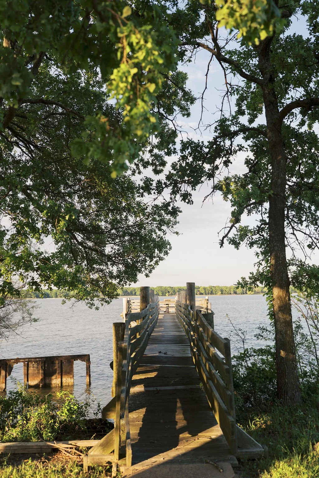 A wooden dock extends over Lake Tawakoni, framed by trees with green leaves on either side, under a partly cloudy sky at Peninsula Ranch in DFW 